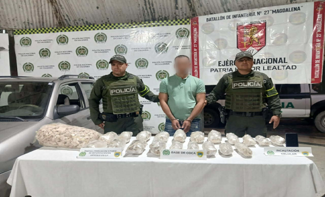 Dos policias con un hombre capturado en la mitad, en frente una mesa con estupefacientes y un auto cologr gris