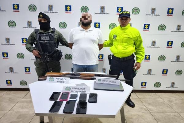 En un espacio institucional, una persona con el rostro difuminado se encuentra de pie entre dos integrantes de la Polic&iacute;a Nacional. Frente a ellos hay una mesa con armas de fuego, varios tel&eacute;fonos celulares incautados y un computador port&aacute;til, debidamente rotulados. Al fondo se observa un respaldo con logos de la Fiscal&iacute;a, la Alcald&iacute;a de Medell&iacute;n y la Polic&iacute;a Nacional