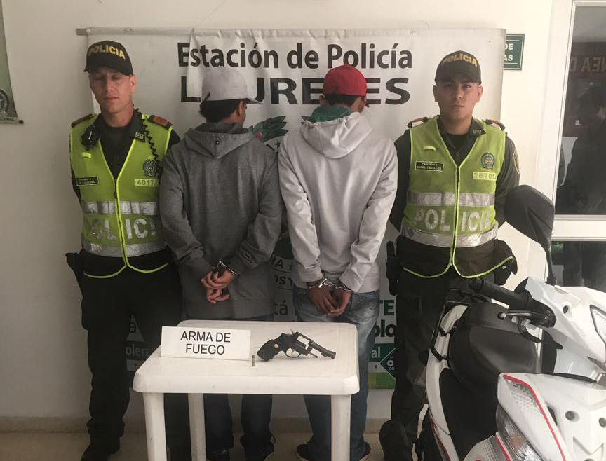 Dos policías custodian dos hombres de espaldas con un arma de fuego incautada en una mesa