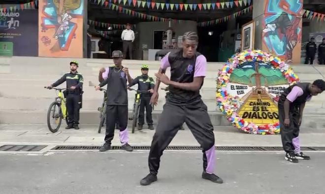 Presentación artística en un espacio público de Medellín: varias personas realizan una danza frente a un escenario, mientras integrantes de la Policía Nacional en bicicleta observan al fondo. Se aprecia decoración colorida y un círculo floral con un mensaje alusivo al diálogo.